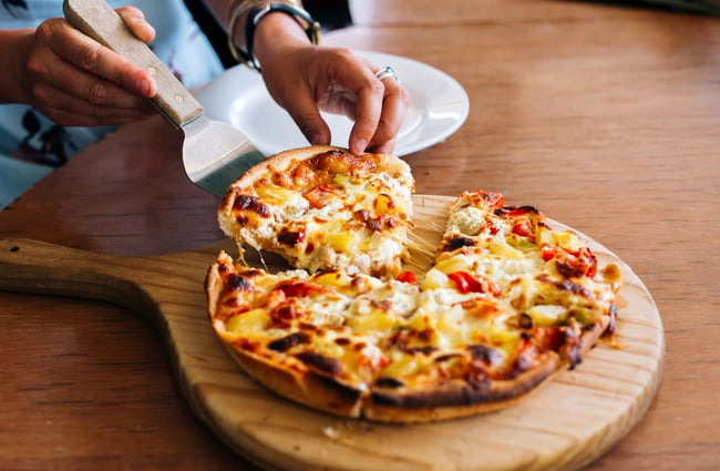 Hands slicing pizza on a board.
