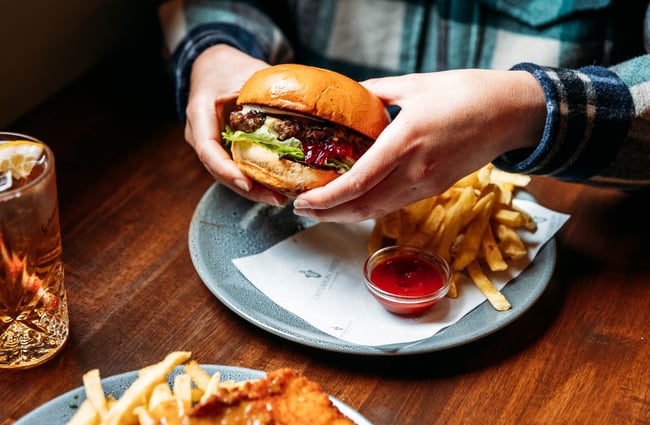 Person holding beef burger up next to a plate of fries and tomato sauce in a pub setting