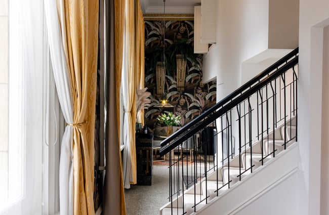 Black handrail on the staircase with yellow curtains and tropical wallpaper in the background at The Eve Apartment.