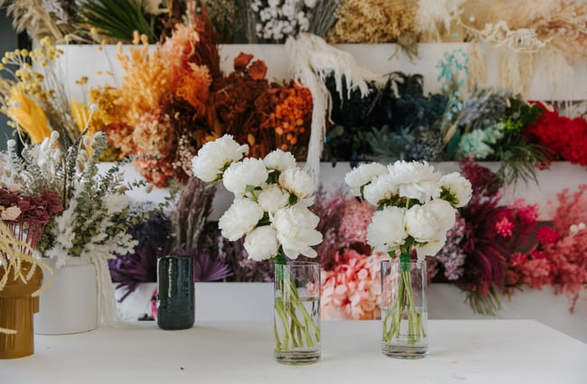 Two glass jars of white flowers with a wall of colourful flowers behind it.