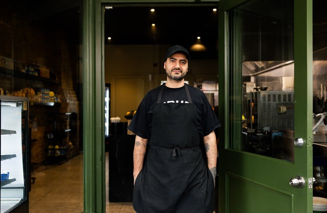 A man wearing an apron standing at the entrance to The Lebanese Grocer.