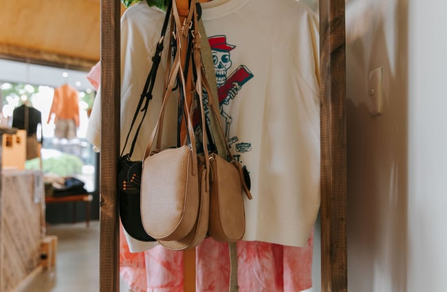 A close up of tan bags sitting on a rack next to white t-shirts at The Red Little Fox store in Fairlie.