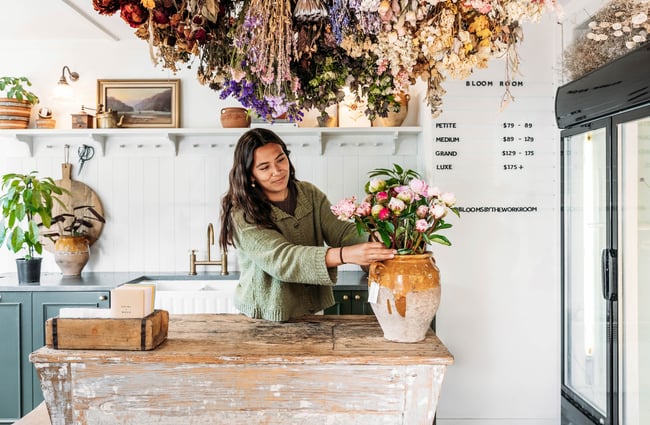 A florist creates a bouquet in a vintage terracotta pot on a weathered wooden counter with drying flowers hung upside down overhead.