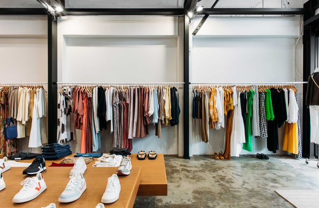 Clothing racks lined up along a big wall and shoes displayed on a big wood table.