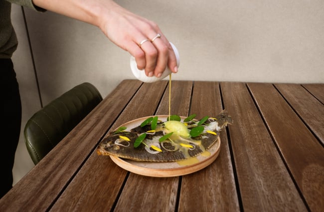 A staff member pours a yellow sauce over a dish of a whole, flat fish.