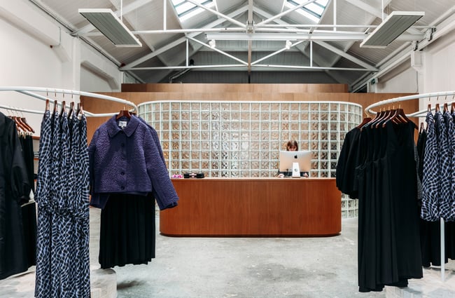Shop assistant behind the counter with clothing racks in front of her in twenty-seven names.