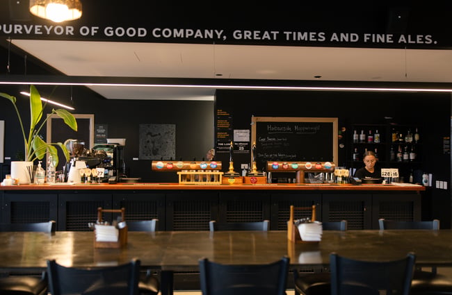 Interior shot of Two Thumb Harbourside with a long table in front of the bar and a woman standing behind cashier