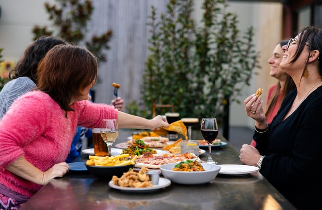 Four friends eating and laughing around a table of drinks and food at Two Thumb Harbourside