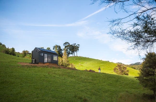 A view of a small black cabin photographed from the bottom of a hill.