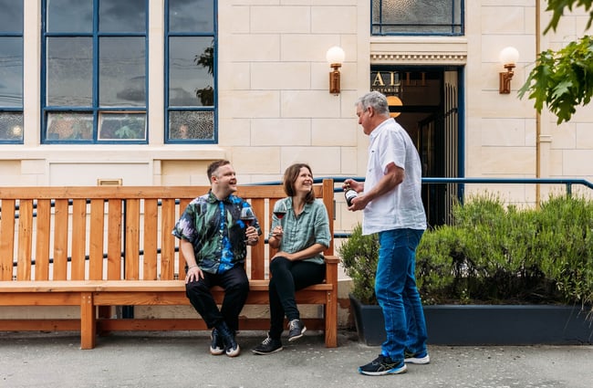 Waiter serving wine to two guests sitting on bench chair outside wine bar