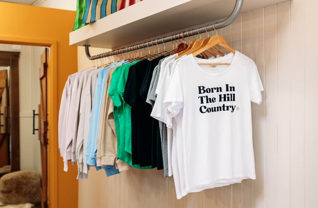 Rack of clothing with a white t-shirt at the front reading "Born In The Hill Country"
