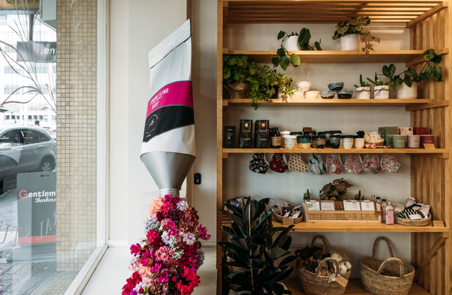 Flowers in the window next to shelves of plants and products on sale.