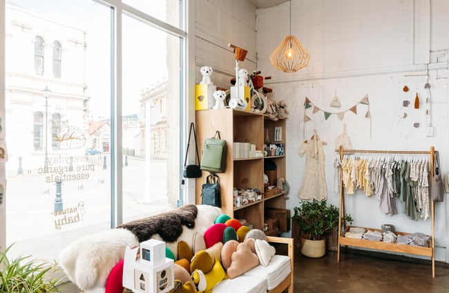 A couch by the window with felted cushions and a sheepskin throw next to a shelf of toys and homeware products and a rack filled with baby clothing