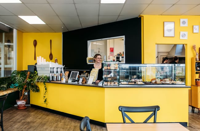 Front counter and food cabinet in the cafe space, with a bright yellow wall behind at Wooden Spoon.