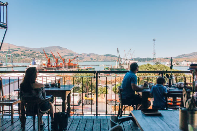People sitting at tables on a balcony looking out towards the Lyttelton Harbour.