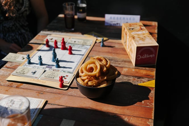 A bowl of onion rings and a board game on a table in the sunshine.