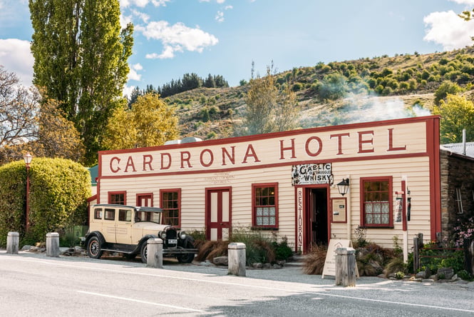 Historic cream and maroon weatherboard frontage of Cardrona Hotel with a vintage cream 1968 Chrysler Model 62 parked in front.