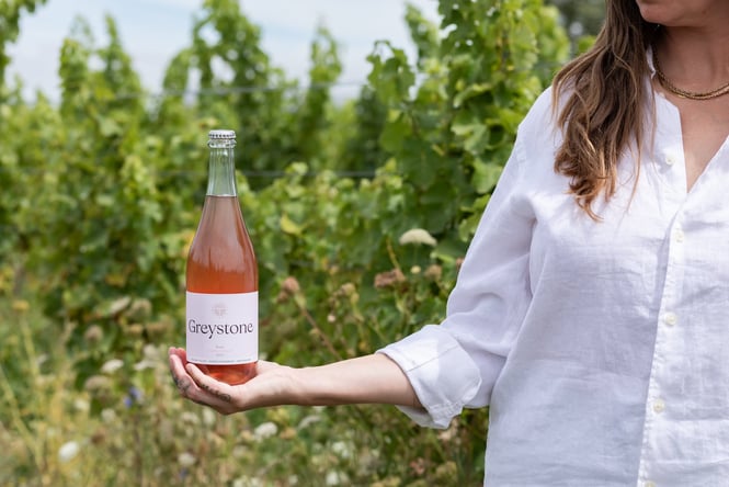 A woman in a white shirt holds a bottle of Greystone Rose Pet Nat against the vineyard's green backdrop at Greystone Wines.