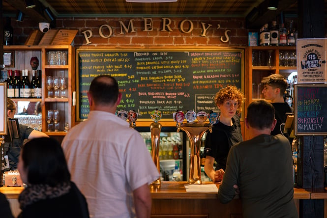 People queuing to order at the bar where on the back wall a big chalkboard lists all of the beers on tap in colourful chalk handwriting.