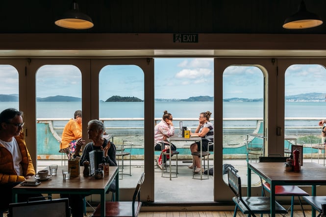 View from inside the first floor Seashore Cabaret cafe out to their deck area where people are dining with a view across the harbour to Wellington city beyond.