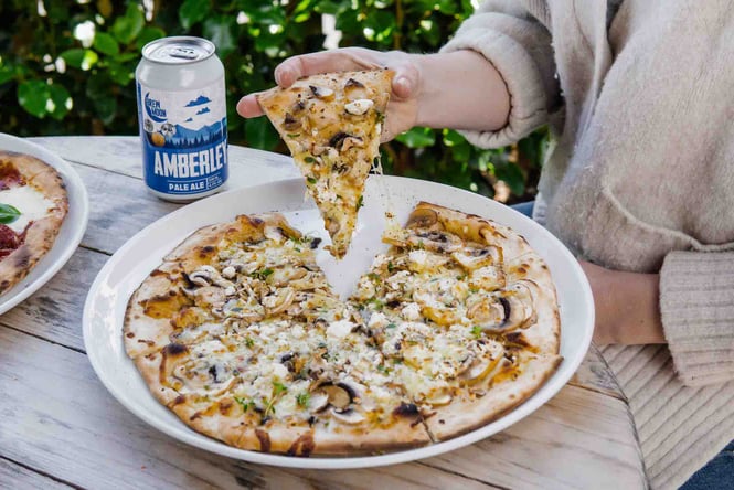 Someone picking up a slice of pizza next to a can of beer at Brew Moon in Amberley
