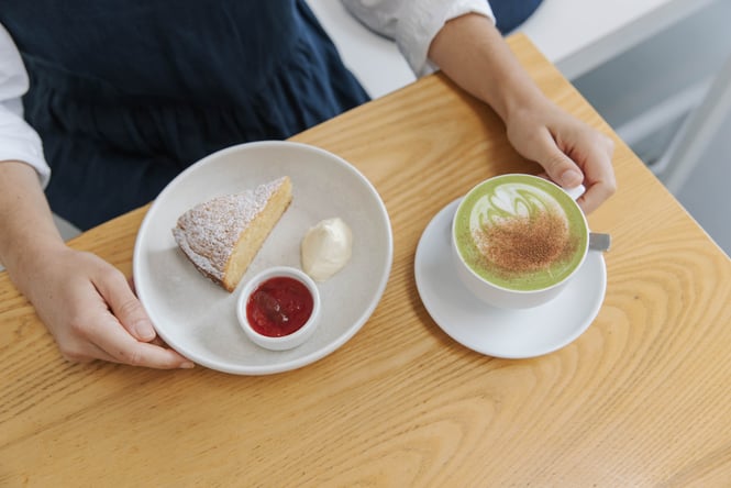 Person sitting at café table with matcha latte and butter cake served with cream and jam