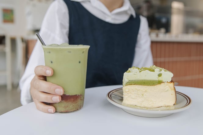 Person sitting at table with an iced matcha strawberry latte next to a piece of matcha basque cheesecake