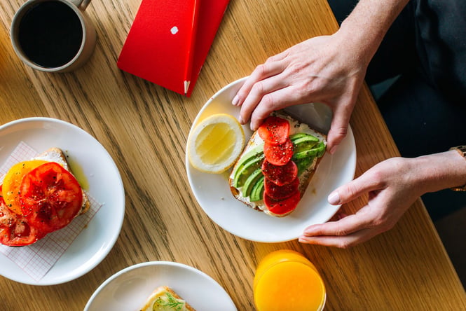 Hands holding a piece of avocado toast at a table inside Customs by Coffee Supreme.
