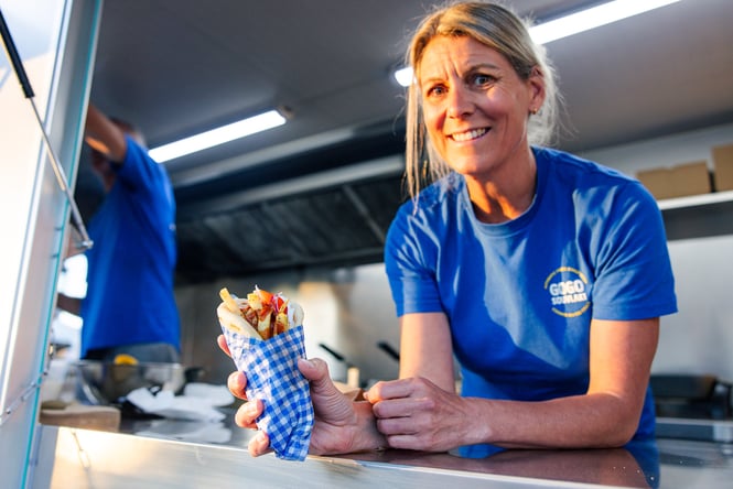 Souvlaki wrapped in blue and white paper being passed to a customer out of a food truck window.