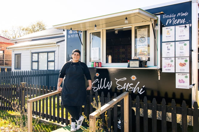 Woman standing in front of a food truck with the menu out front and a house in the background.