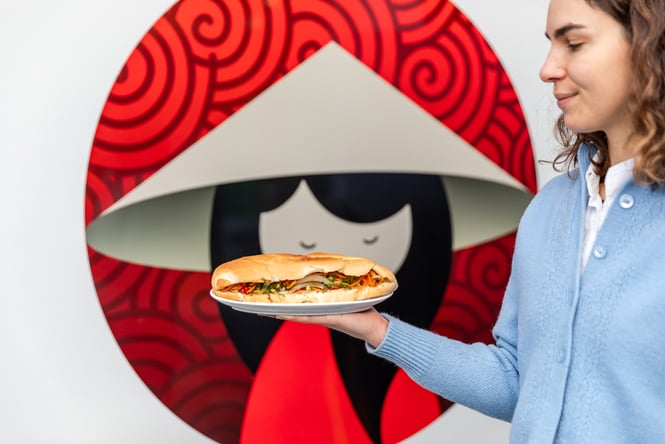 Person holding up a plate with a fresh banh mi in front of Miss Saigon logo