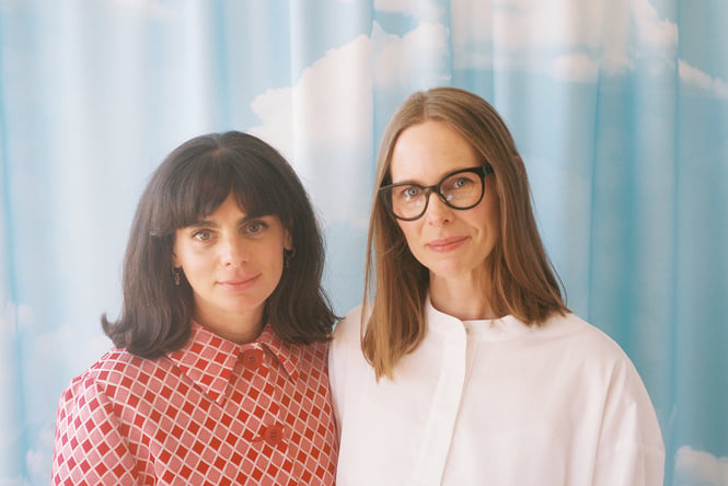 Two women standing together in front of a printed curtain.