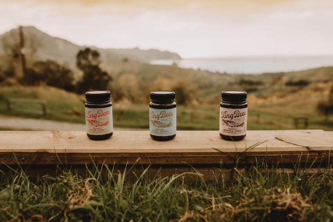 Three jars of King Bees Honey are lined up on a wooden fence post with rolling hills and the bay in the background.
