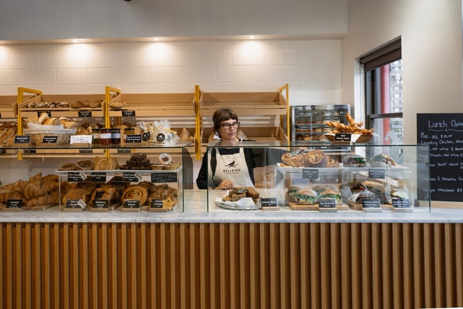 Woman in a Bellbird apron standing behind cabinet of baked goods including sandwiches, pastries and slices