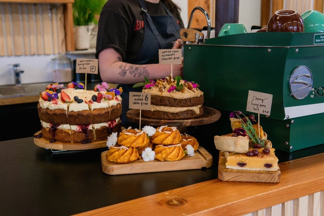 Beautiful cakes and tarts topped with flowers and fruits on a cafe counter with a woman making coffee in the background