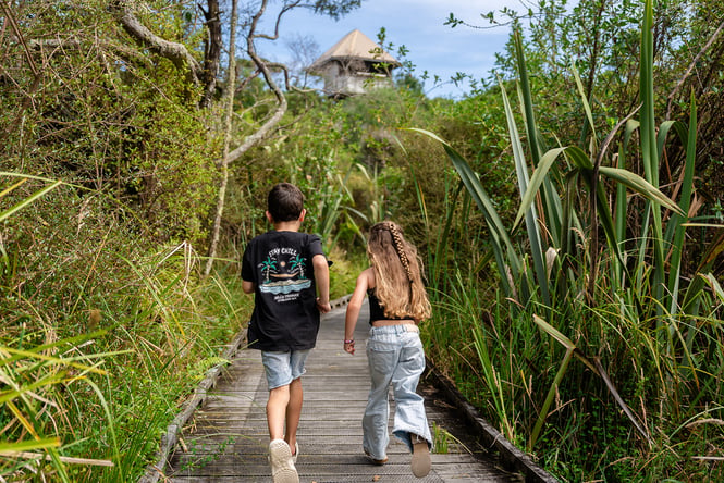 Two kids running on a boardwalk through native bush.