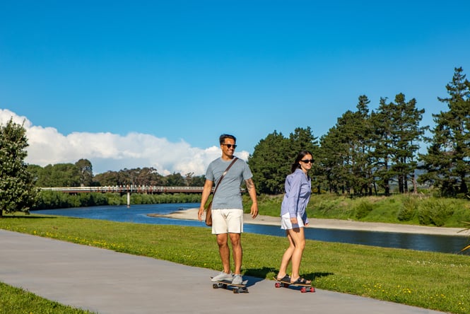 A couple are skateboarding on the pathway next to the river.