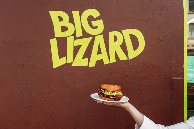 Someone holding a fried chicken burger in front of Big Lizard sign in Dunedin