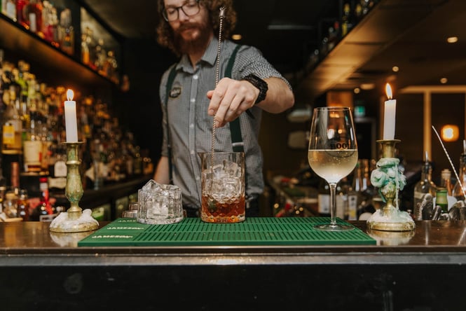 Bartending stirring cocktail on bar counter next to a glass of wine