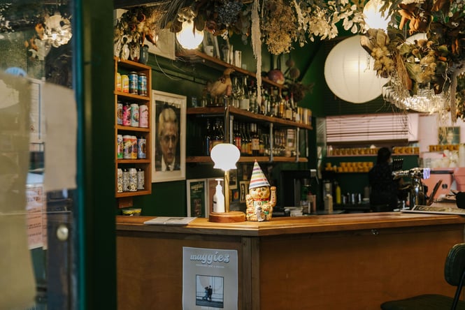 Looking through window into Maggies bar and cafe in Dunedin with dried floral arrangement hanging from ceiling and a lucky fortune sitting on counter with a hat on