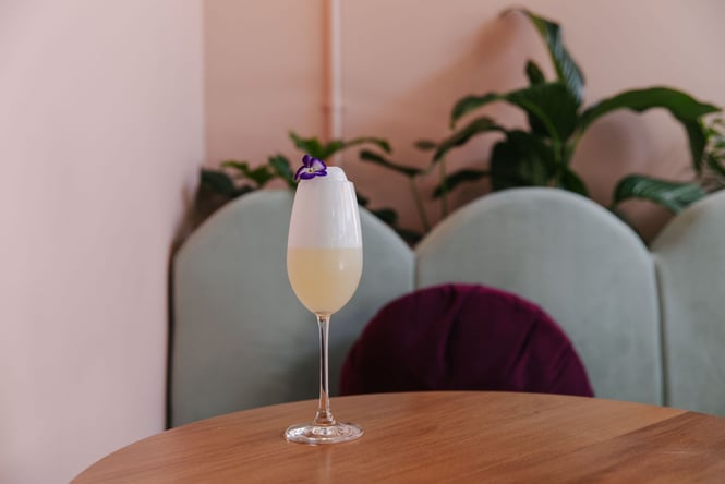 Close up of citrusy cocktail on a table at Gin Gin with a foamy top and a purple pansy garnish