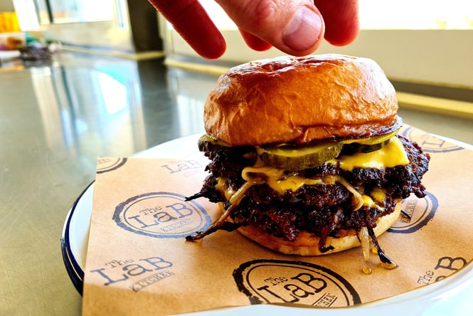 A hand over top of a burger on a table.