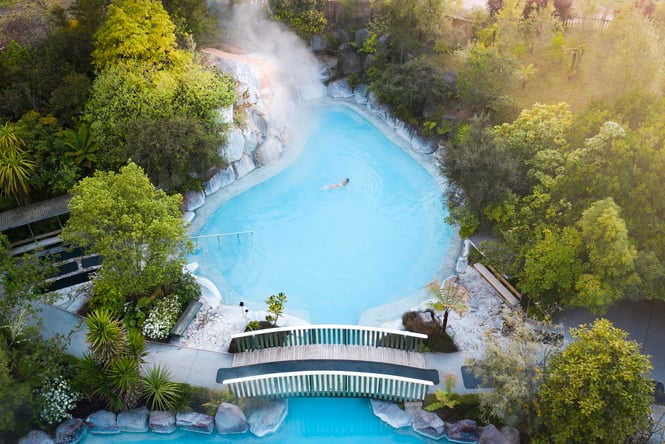 Bird’s eye view of a bright blue steaming hot springs with a bridge walkway going over the pool.