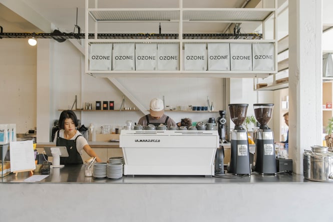 Two baristas standing behind counter at Ozone coffee with shelves of coffee bags overhead