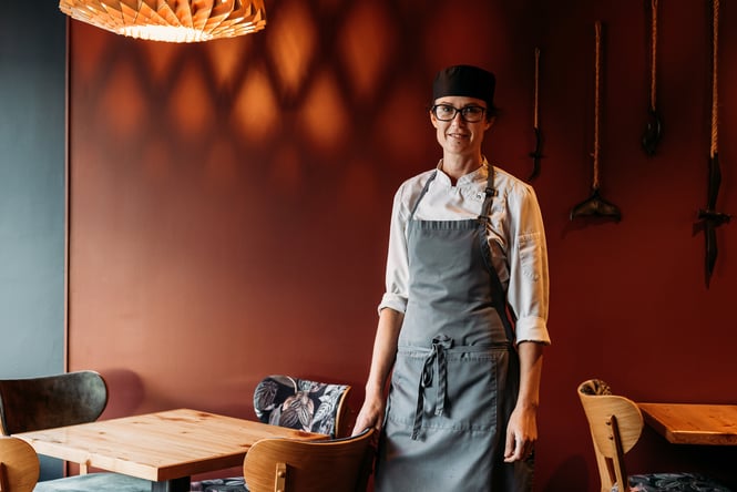 A female chef smiles at the camera from the dining area of a restaurant.