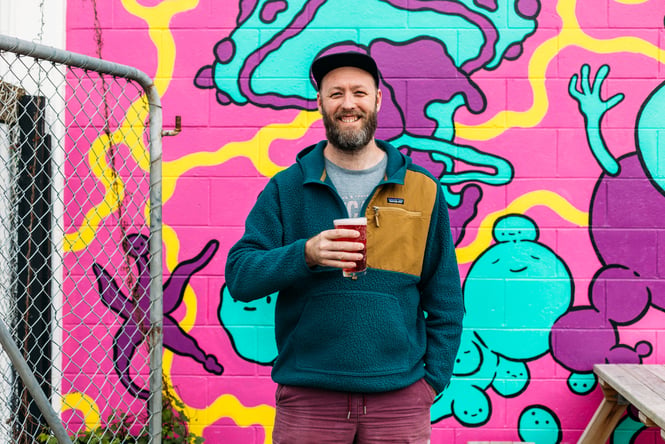 A man holding a beer smiling to camera in front of a bright wall mural.