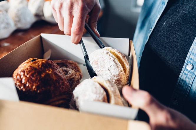 A person using tongs to dish donuts into a take away box.