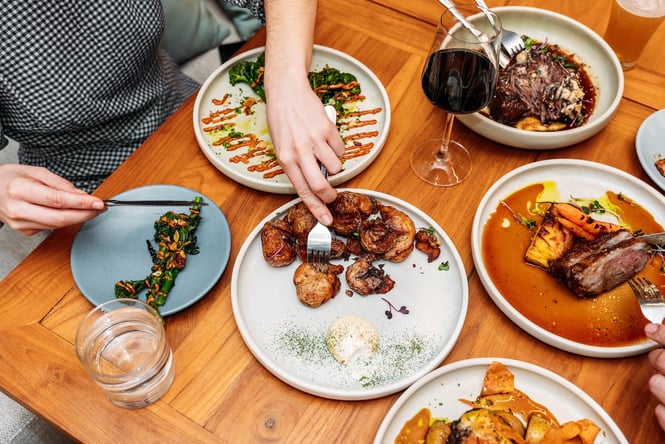 Two people serving themselves from plates of savoury dishes spread across a wooden table.