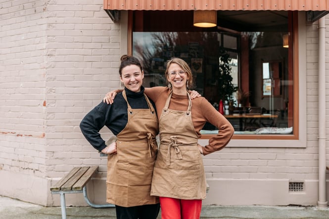Two women smiling to camera outside their cafe.