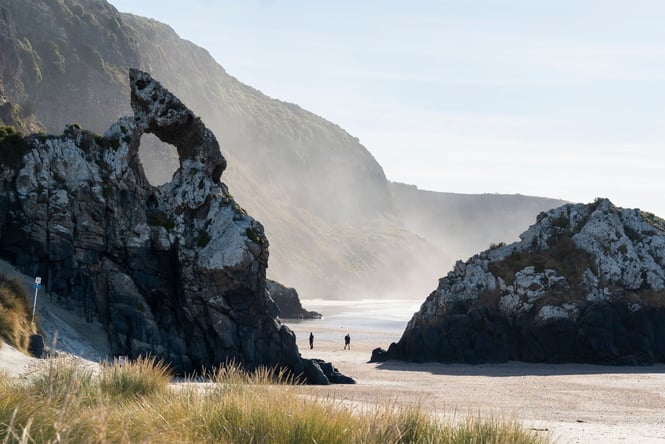Two people walking between two giant rocks on Aramoana Beach.